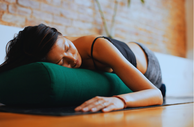 A woman rests on her side on a yoga mat, eyes closed and head supported by a bolster, in a quiet restorative yoga pose.